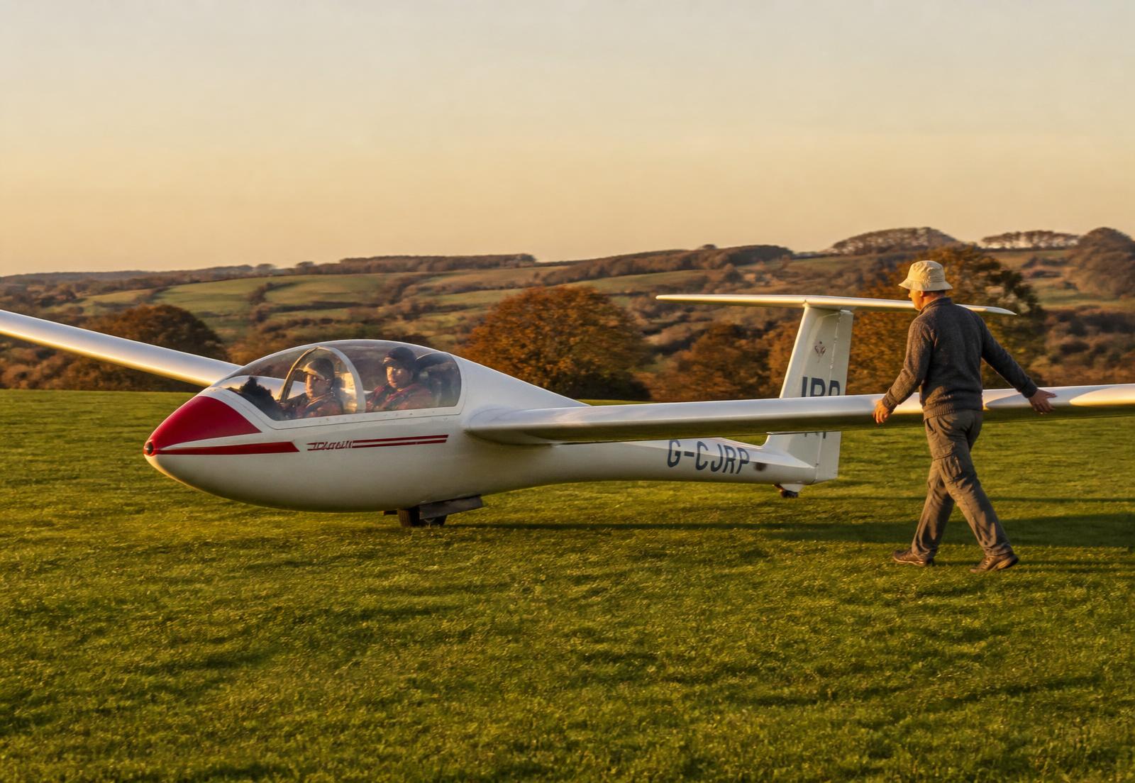 A two seat training glider on the grass at Vale Airfield with a wing runner alongside in evening light